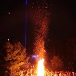 A crowd around a bonfire with blue laser going up to sky