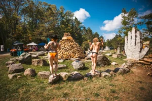 HARVEST 2025, @milabright_photo-3481 Two women in bikini bottoms stand near a teepee-like unlit wood bonfire