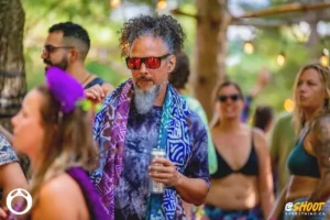 man-by-zac-mac. Man with curly hair and sunglasses at an outdoor festival, holding a drink